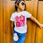 Girl wearing sunglasses and a pink headband, in a white T‑shirt with Powerpuff Girls graphics, leaning against a wooden wall.
